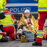 Notfallsanitäter versorgen einen verletzten Menschen mit Kopfverband und Sauerstoffmaske vor einem Polizeistreifenwagen und Notfallausrüstung