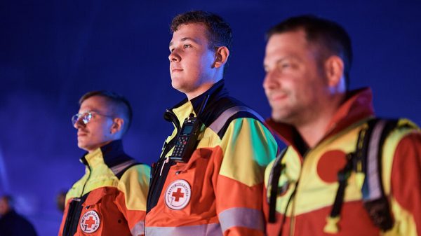 Notfallreform: Drei Einsatzkräfte des DRK-Rettungsdienstes in orange-gelber Warnkleidung vor blauem Hintergrund