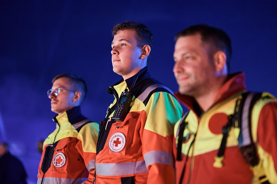 Notfallreform: Drei Einsatzkräfte des DRK-Rettungsdienstes in orange-gelber Warnkleidung vor blauem Hintergrund